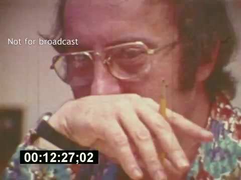The still from 'Sixty Second Spot' captures a contemplative moment in the creation of a 7-Up commercial from the mid-1970s. The focus is on a man, possibly a creative professional, wearing glasses and a colorful shirt. He holds a pencil thoughtfully against his mouth, suggesting deep consideration or brainstorming. The frame includes a timecode, indicating it's taken from an edit and is marked “Not for broadcast,” emphasizing its behind-the-scenes context in the filmmaking process. This image reflects the collaborative effort involved in commercial production.