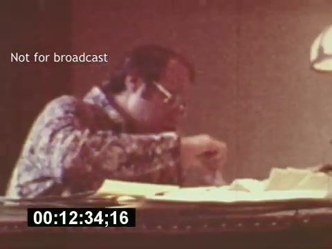 The still from 'Sixty Second Spot' features a man in a patterned shirt, focused on working at a desk cluttered with papers. It captures a behind-the-scenes moment during the creative process of developing a 7-Up commercial, showcasing scriptwriting and preparation stages from the mid-1970s. The annotation “Not for broadcast” indicates it’s a production still rather than a final cut.