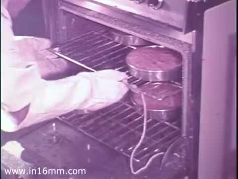 The still from the 1975 American Gas Association PSA shows a person in protective gloves and clothing handling two cake pans inside an oven. The scene emphasizes safety and proper appliance use, likely illustrating the importance of testing gas appliances to ensure safety and efficacy. The overall tone suggests a focus on responsible appliance management.