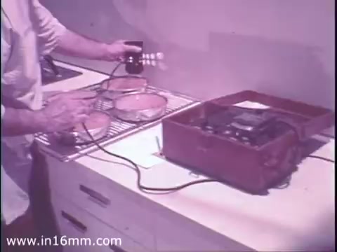 The still from the 1975 'American Gas Association PSA' shows a person testing gas appliances in a kitchen setting. The individual is using a device connected to a red testing unit, likely assessing the safety or functionality of the appliances on a cooling rack. The visual emphasizes the importance of appliance testing for safety, aligning with the PSA's message.