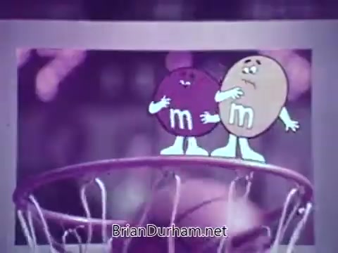 The still from the M&M's animated basketball commercial features two anthropomorphic M&M candies, one purple and one tan. They are positioned near the basketball hoop, looking towards the action, likely reacting to a game scenario. The playful nature of the characters adds a humorous touch to the vibrant, animated scene.