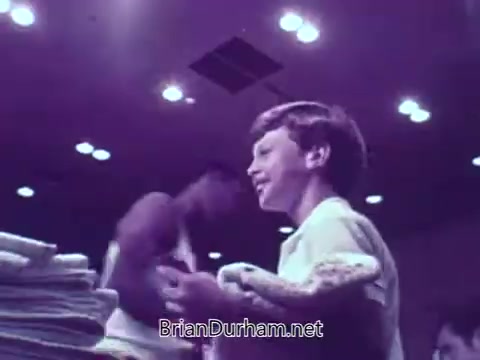 The still from the M&M's animated basketball commercial features a young boy smiling enthusiastically, holding a towel. In the background, there are two figures, likely basketball players, engaged in activity. The lighting is bright, emphasizing the playful and energetic atmosphere typical of M&M's advertising. The focus is on fun and excitement, capturing the joy associated with both basketball and the candy.