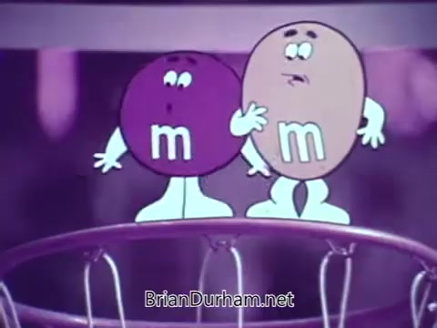 The still features two animated M&M's characters, one purple and one tan, standing near a basketball hoop. They appear to be engaged in a lighthearted moment, likely discussing the game. The scene is colorful and playful, characteristic of M&M's commercials, emphasizing fun and camaraderie among the candy characters.