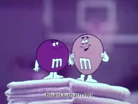 The still features two animated M&M's candies, one purple and one brown, appearing cheerful and friendly. They are likely standing on a stack of towels, set against a background that hints at a basketball setting with a hoop. The playful interaction emphasizes their personalities, typical of M&M's commercials that combine humor and approachability.