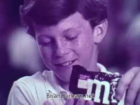 The still features a young boy with freckles, smiling as he holds a package of M&M's, possibly in a basketball-themed context. The commercial likely showcases the fun and enjoyment of M&M's, emphasizing their appeal during a sporting event. The colors and animation style contribute to a playful atmosphere, typical of M&M's advertising.