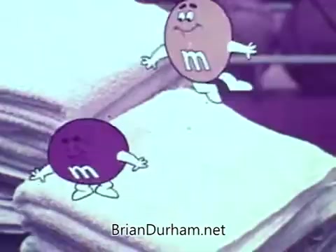 The still features animated M&M's characters, specifically a purple and a brown candy, set against a backdrop of soft fabric. The characters are depicted with expressive faces and playful postures, highlighting their vibrant personalities. This scene is part of a basketball-themed commercial, emphasizing fun and humor in the brand's marketing approach.