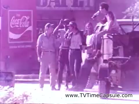The still features a group of young adults, gathered around a vehicle, enjoying each other's company and drinks. They appear joyful and relaxed, embodying a spirit of camaraderie. In the background, a vintage Coca-Cola sign promotes the brand, reinforcing the theme of happiness associated with sharing a Coke. The scene is nostalgic, reflecting a carefree moment outdoors.