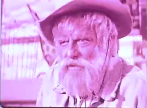 The still features an older man with a long beard and wearing a wide-brimmed hat, likely portraying a rugged character. The image has a distinct pink tint, suggesting it’s from a 16mm print that may have faded over time. The setting appears to be western-themed, aligning with the film's context, 'Scandalous John.' The man's expression is thoughtful, indicating possible depth in his character's storyline.