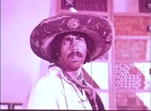 The still from 'Scandalous John' features a man wearing a large, intricately decorated sombrero. He has a serious expression, with a mustache and visible facial features indicating concern or intensity. The background suggests a rustic setting, with elements of wood and patterned decor. The overall color tone appears warm, likely due to the 16mm film's characteristics. The scene evokes a sense of drama and captures a moment of tension.