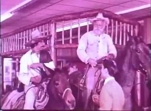 The still from 'Scandalous John' features two characters on horseback in an indoor setting. One man, dressed in light clothing with a cowboy hat, sits on his horse while another man, an older character in a light-colored outfit and white hat, stands beside him. The background includes wooden beams and elements indicative of a rustic environment. The overall color tone has a pinkish hue, typical of vintage 16mm prints.
