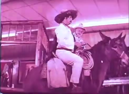 The still from 'Scandalous John' features a scene in which a man wearing a sombrero and traditional attire is seated on a horse inside what appears to be a building, possibly a saloon or similar venue. Another character, also dressed in Western attire, is nearby. The color palette has a pinkish hue, characteristic of certain film prints, adding a stylized effect to the scene. The setting suggests a humorous or lighthearted context typical of the film's theme.