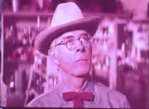 The still features a character wearing a light-colored cowboy hat and round glasses, with a distinctive red bow tie against a checkered shirt. The color palette appears slightly faded, typical of a 16mm print, with a soft focus that evokes a vintage feel. The background suggests a lively setting, possibly a rural store or similar environment, conveying a whimsical or comedic tone in line with the film's themes.