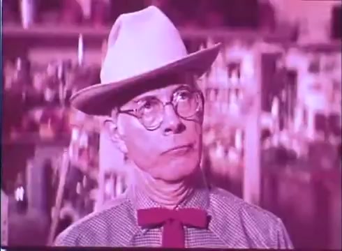 The still features an older man with glasses, a bowtie, and a white hat, suggesting a whimsical or quirky character. His expression is thoughtful or curious, set against a blurred background that hints at a shop or market environment. The vibrant colors typical of a 16mm print highlight the film's nostalgic and lighthearted tone, aligning with the comedic narrative of 'Scandalous John.'