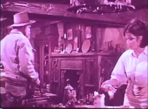 The still from 'Scandalous John' features two characters in a rustic interior setting. On the left, a man in a hat and western attire stands, appearing to engage with the scene. To the right, a woman in a white blouse is busy with a task, perhaps preparing food or organizing items on a table. The warm tones and vintage look of the 16mm print enhance the nostalgic atmosphere of the film. The decor includes wooden furniture and various objects that contribute to the Western feel.