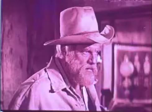 The still from 'Scandalous John' features a rugged character, likely the protagonist, portrayed by Brian Keith. He has a weathered face, a full beard, and is wearing a wide-brimmed hat. The lighting casts a dramatic tone, enhancing the character's rough demeanor. The background hints at a rustic setting, typical of western films, with elements that suggest a historical or frontier atmosphere. Overall, the image captures a tense or thoughtful moment.