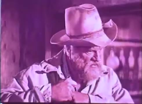 The still from 'Scandalous John' features a rugged man with a bushy beard, wearing a dusty cowboy hat and a light-colored jacket. His expression is gruff, suggesting a moment of contemplation or tension. The background is slightly blurred, with indistinct shapes that hint at a rustic setting, likely emphasizing the Western theme of the film. The color tone appears warm, typical of vintage film prints.