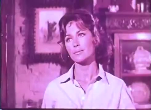 The still from 'Scandalous John' features a woman with shoulder-length hair, looking thoughtfully or wistfully. The color palette has a vintage hue, typical of 16mm film, and hints at a warm, nostalgic atmosphere. The background includes decorative elements, suggesting an intimate indoor setting. Her expression conveys a sense of contemplation or introspection.