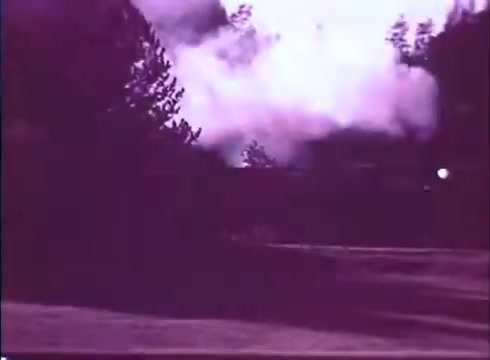 The still from 'Scandalous John' depicts a smoky scene, possibly from an explosion or fire, set against a backdrop of trees. The coloring has a purple tint, and the image captures a moment of dramatic action, suggesting tension or conflict in the film. The framing emphasizes the surrounding natural environment, hinting at an outdoor setting.