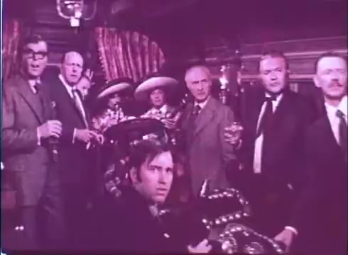 The still from 'Scandalous John' features a group of men in formal attire, some holding drinks, gathered in a richly decorated interior. The mood appears tense or dramatic, with several individuals looking intently at the camera. The setting suggests a social gathering with an air of intrigue, characteristic of the film's themes. The color tone is slightly muted, typical of older 16mm prints.
