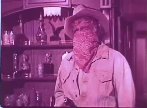 The still from 'Scandalous John' features a rugged character dressed in a cowboy outfit. He wears a wide-brimmed hat and a red bandana that covers part of his face. The background suggests a Western setting, with shelves displaying glass bottles, possibly indicating a saloon or a rustic interior. The overall color tone appears warm, lending a vintage feel typical of 16mm prints. The character's posture and attire evoke a sense of intrigue and tension.