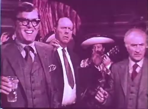 The still from 'Scandalous John' features a group of men in a lively setting. The central figure is smiling broadly, wearing glasses and holding a glass, suggesting a festive atmosphere. Surrounding him are two other men; one appears surprised or concerned, while another looks serious. In the background, a musician can be seen with a guitar, hinting at a celebratory or comedic scene typical of the film's lighthearted tone. The color tone is warm and slightly faded, characteristic of a 16mm print.