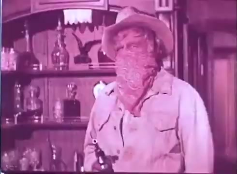 The still from 'Scandalous John' features a man dressed in western attire, including a hat and a bandana covering the lower half of his face. He holds a firearm, standing in an interior space that appears to be a bar or saloon, with various bottles displayed on shelves behind him. The overall atmosphere suggests a tense or dramatic moment typical of the western genre. The color tone indicates it is a vintage 16mm print.
