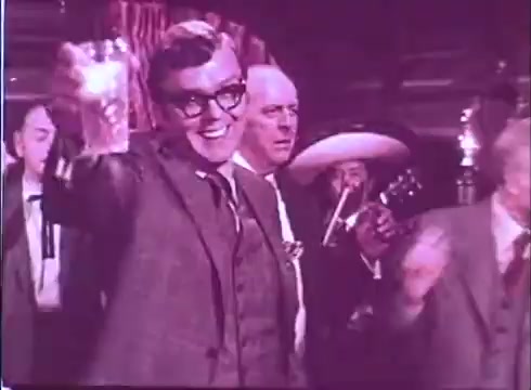 The still from 'Scandalous John' features a gathering where a man in glasses raises a glass, smiling cheerfully. The scene is lively with others in the background, including a man wearing a sombrero and another holding a guitar. The color tone appears faded, typical of a 16mm print, contributing to a nostalgic atmosphere. The overall mood suggests celebration or camaraderie.