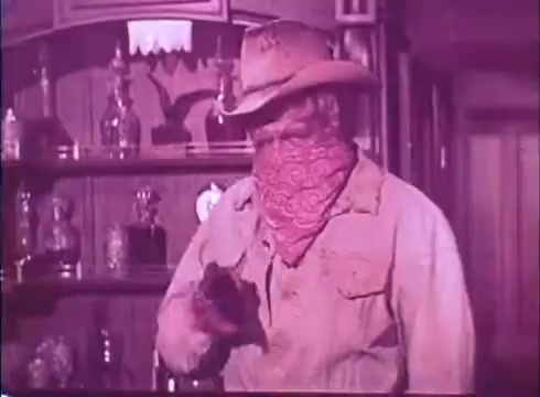 The still features a cowboy character wearing a wide-brimmed hat and a red bandana covering his face, suggesting a classic Western theme. He stands in an interior setting, likely a saloon, with shelves displaying glass bottles behind him. The atmosphere appears vibrant, with a heightened color palette typical of 16mm film prints. The cowboy is poised with a firearm, adding tension to the scene.