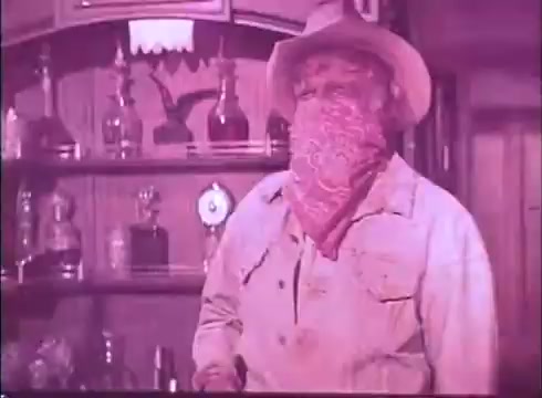 The still from 'Scandalous John' features a character portrayed by Brian Keith, wearing a cowboy hat and a red bandana over his face. He stands in a western-themed setting, possibly a bar, with various bottles and trophies visible behind him. The overall tone of the image has a vintage appearance due to the 16mm format. The character's expression is obscured by the bandana, adding a sense of intrigue.