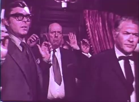 The still from 'Scandalous John' features three men, each with their hands raised in a gesture of surrender or compliance. The man on the left wears glasses and a suit, displaying a tense expression. The central figure is older and looks startled, while the man on the right, in a tuxedo, appears serious. The background suggests a formal setting, accentuated by draped curtains. The color and grain typical of a 16mm print give it a vintage feel. Overall, the scene conveys a moment of tension or conflict.