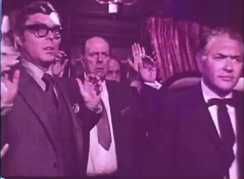 The still from 'Scandalous John' features three men with their hands raised, suggesting a moment of tension or surprise. The figures are dressed in 1970s attire, with the man on the left wearing glasses and a suit, indicative of the era. The background is dimly lit, enhancing the dramatic atmosphere. The overall composition implies a pivotal scene within the film's narrative, emphasizing the characters' reactions.