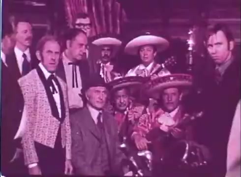 The still from 'Scandalous John' features a group of men posed together in a colorful setting. The foreground includes a man in a stylish suit with a patterned jacket and a cowboy hat, surrounded by other men, some wearing traditional Mexican attire with sombreros. In the background, musicians are seen holding instruments, creating a festive atmosphere. The overall tone of the image reflects the comedic and lively essence of the film. The print has a vintage quality typical of 16mm film formats.