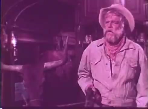 The still from 'Scandalous John' features Brian Keith as an older, rugged character in a western setting. He wears a cowboy hat and a light-colored shirt, paired with a bandana around his neck. The scene is dimly lit with a nostalgic, vintage tone, highlighting Western elements like rustic decor and a bull's head in the background. Keith's expression conveys a sense of concern or contemplation, fitting the film's comedic and adventurous themes. The overall mood captures a blend of humor and western nostalgia.