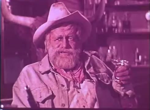 The still from 'Scandalous John' features an older man with a bushy beard, wearing a cowboy hat and a denim jacket. He holds a glass in his hand, likely containing a drink, while sitting in a rustic setting reminiscent of a bar. The color tone appears to be slightly faded, typical of a 16mm print, and captures a laid-back, Western atmosphere. The character expresses a rough, jovial demeanor that fits the film's promotional style.