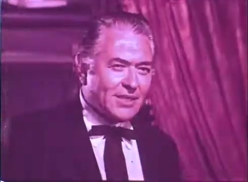 The still from 'Scandalous John' features a middle-aged man dressed in formal attire, including a tuxedo and bow tie. He has slicked-back hair and a confident smile, suggesting charm and charisma. The background is softly lit with warm tones, enhancing the theatrical atmosphere of the scene. The image has a vintage quality typical of 16mm film prints.