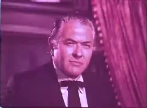 The still from 'Scandalous John' features a serious-looking male character portrayed by Brian Keith. He is dressed in formal attire, including a bow tie, and is positioned in front of a rich, textured background, likely indicating a luxurious or dramatic setting. The color tone of the image has a vintage quality, typical of a 16mm print, imparting a nostalgic feel to the scene.