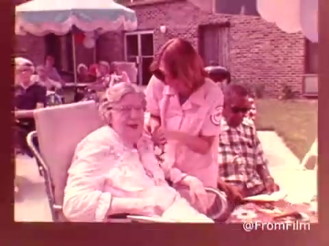 The still from the 1970s American Health Care Association commercial features a cheerful scene in a care facility. An elderly woman sits in a wheelchair, smiling as a caregiver assists her. In the background, a small group of residents enjoys the outdoors, with brick buildings and shaded areas visible. The atmosphere appears warm and welcoming, highlighting community and care for elderly individuals.   .