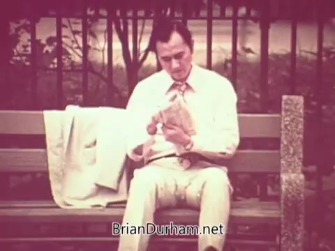 The still depicts a man in a light-colored suit sitting on a park bench, looking at a folded newspaper. His expression appears contemplative or concerned, suggesting he may be pondering job opportunities. A lightweight jacket rests beside him, and the setting is serene, characterized by muted colors typical of 16mm film. The atmosphere conveys a sense of introspection related to employment or job searching.