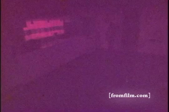 The still from 'Formica Floor Shine' captures a dimly lit scene with a heavy purple tint, likely due to film deterioration or specific processing techniques typical of 16mm prints. The image suggests an interior space, possibly with furniture or surfaces that appear abstracted and undefined in the low lighting. The overall mood is atmospheric, evoking a sense of nostalgia or reflection.