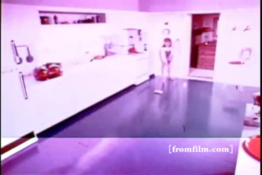 The still from 'Formica Floor Shine' features a brightly lit domestic kitchen with a shiny, polished floor. A child stands in the center, likely engaged in play or household activity. The overall aesthetic is vivid, showcasing pastel colors, particularly a pink hue that dominates the scene. The composition emphasizes a clean, modern space typical of mid-20th century design, reflecting leisure and domesticity.