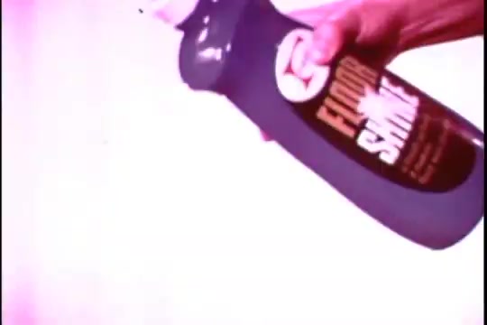 The still features a hand holding a bottle of 'Floor Shine,' a cleaning product. The label is prominently displayed, emphasizing its purpose. The color palette is muted, typical of 16mm film, with a slight graininess, giving it a vintage feel. The background is light, allowing the product to stand out as the focal point.