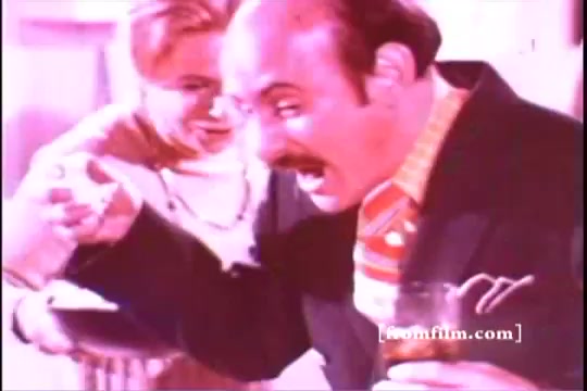The still from 'Formica Floor Shine' features a man with a mustache, animatedly gesturing while holding a drink in one hand. A woman appears to be serving him something. The color palette is warm, enhancing the lively domestic atmosphere. The scene suggests a moment of excitement or humor in a social setting.