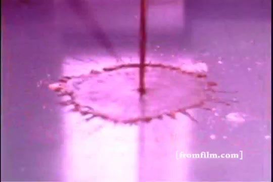 The still from 'Formica Floor Shine' depicts a liquid being poured onto a shiny surface, creating ripples and splashes. The scene emphasizes the reflective quality of the surface, enhanced by a pink hue. The focus on the liquid's interaction with the floor evokes themes of cleanliness and surface care, characteristic of the film's exploration of mundane objects and their aesthetic.