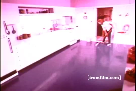 The still from 'Formica Floor Shine' features a brightly lit kitchen with a shiny, wet floor. A person, dressed in a white shirt, is seen mopping the floor, emphasizing cleanliness and maintenance. The overall color palette is warm, with pink tones, highlighting a nostalgic domestic scene. The kitchen appears modern for its time, showcasing a focus on domestic aesthetics and practical care for home spaces.