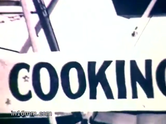 The still features a prominently displayed sign that reads 'COOKING,' likely part of a cooking competition at a county fair. The context suggests a nostalgic reflection on the tradition of awarding prizes for cooking, with a specific emphasis on dishes made using Mueller's egg noodles. The voiceover highlights the special quality of the noodles, implying they contribute to the success of many recipes and the popularity of the brand.