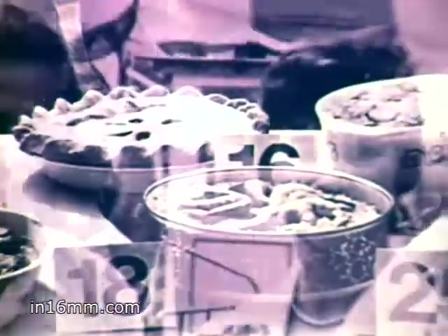 The still features a display of various dishes at a county fair, showcasing the competitive spirit of cooking. Prominent among the items are pastries and desserts, numbered for judging. The voiceover highlights the long-standing tradition of awarding cooking prizes and hints at the popularity of Mueller's egg noodles in award-winning recipes. The overall atmosphere conveys a sense of community and culinary pride.