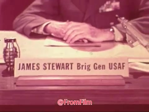 The still from the Civil Air Patrol PSA features a close-up of a table with a nameplate reading 'JAMES STEWART Brig Gen USAF.' In the scene, James Stewart, a prominent actor and Brigadier General in the U.S. Air Force, is likely addressing the audience in a position of authority. The setting suggests a formal presentation or announcement related to the Civil Air Patrol. Various objects, including what appears to be a grenade, indicate a military context.