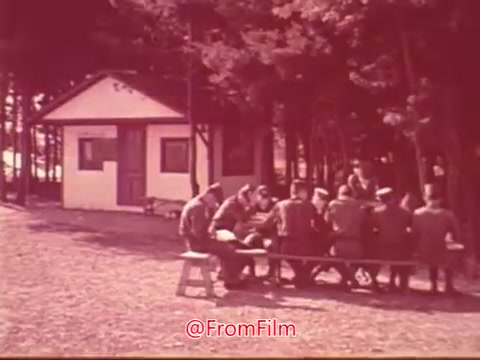 The still from the 'Civil Air Patrol PSA with Jimmy Stewart' depicts a group of individuals seated around a picnic table in front of a small building surrounded by trees. They appear to be engaged in conversation, possibly discussing their activities or coordinating efforts related to the Civil Air Patrol. The overall ambiance suggests a community-focused environment, emphasizing camaraderie and teamwork.