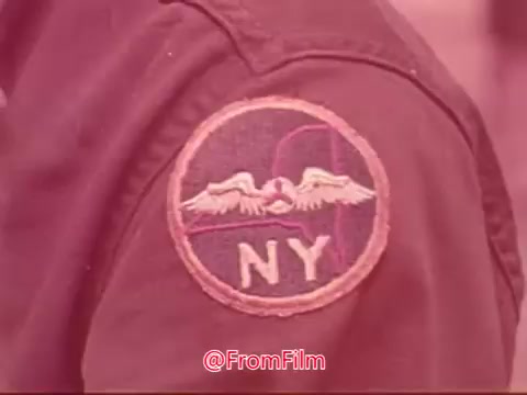 The still features a close-up of a jacket patch displaying the emblem of the Civil Air Patrol, specifically highlighting the letters 'NY,' indicating the New York group. The patch includes wings and other design elements typical of the organization's insignia. The image is tinted in a pink hue, characteristic of 16mm film.