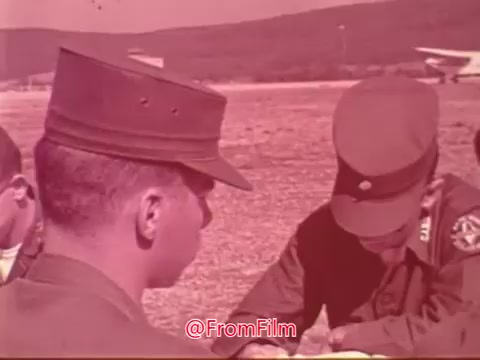 The still from the 'Civil Air Patrol PSA with James Stewart' showcases a group of uniformed individuals engaged in a discussion, likely involving aviation or training matters. The setting appears to be outdoors, possibly at an airfield, with aircraft visible in the background. The focus on the characters suggests a serious demeanor, reflecting the purpose of the Civil Air Patrol in training and community service. The colors and grain typical of 16mm film enhance the vintage feel of the image.